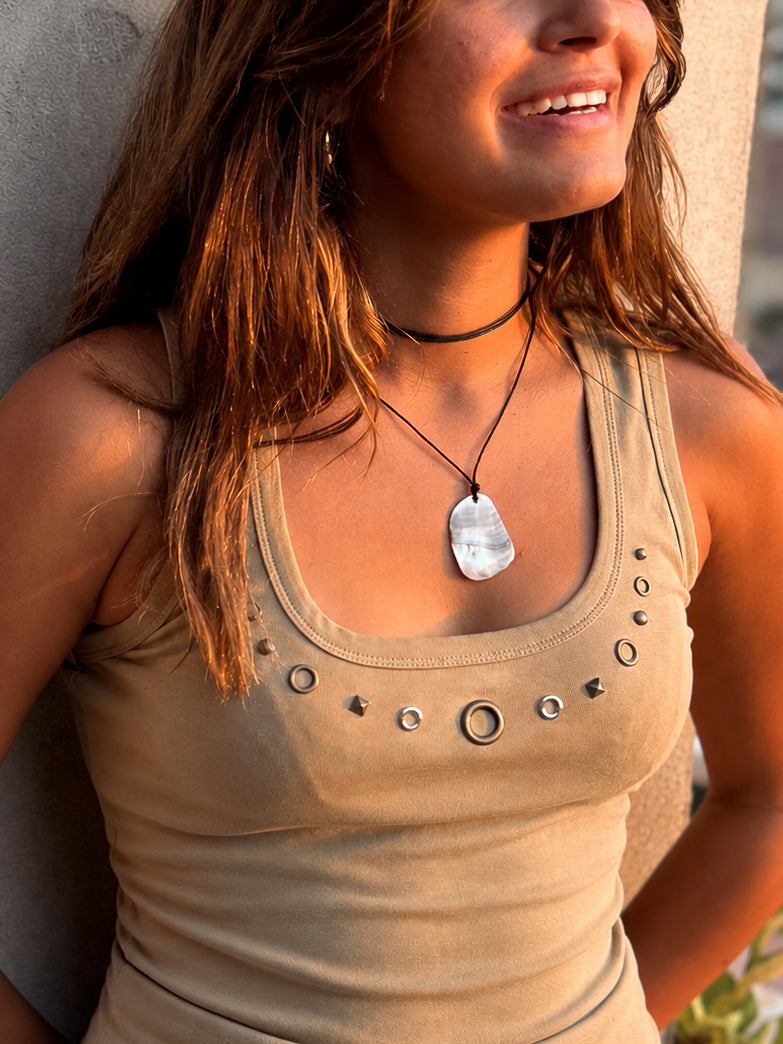 Woman wearing a beige tank top with a necklace featuring a large stone pendant tied to a black wax cord, standing against a neutral background.