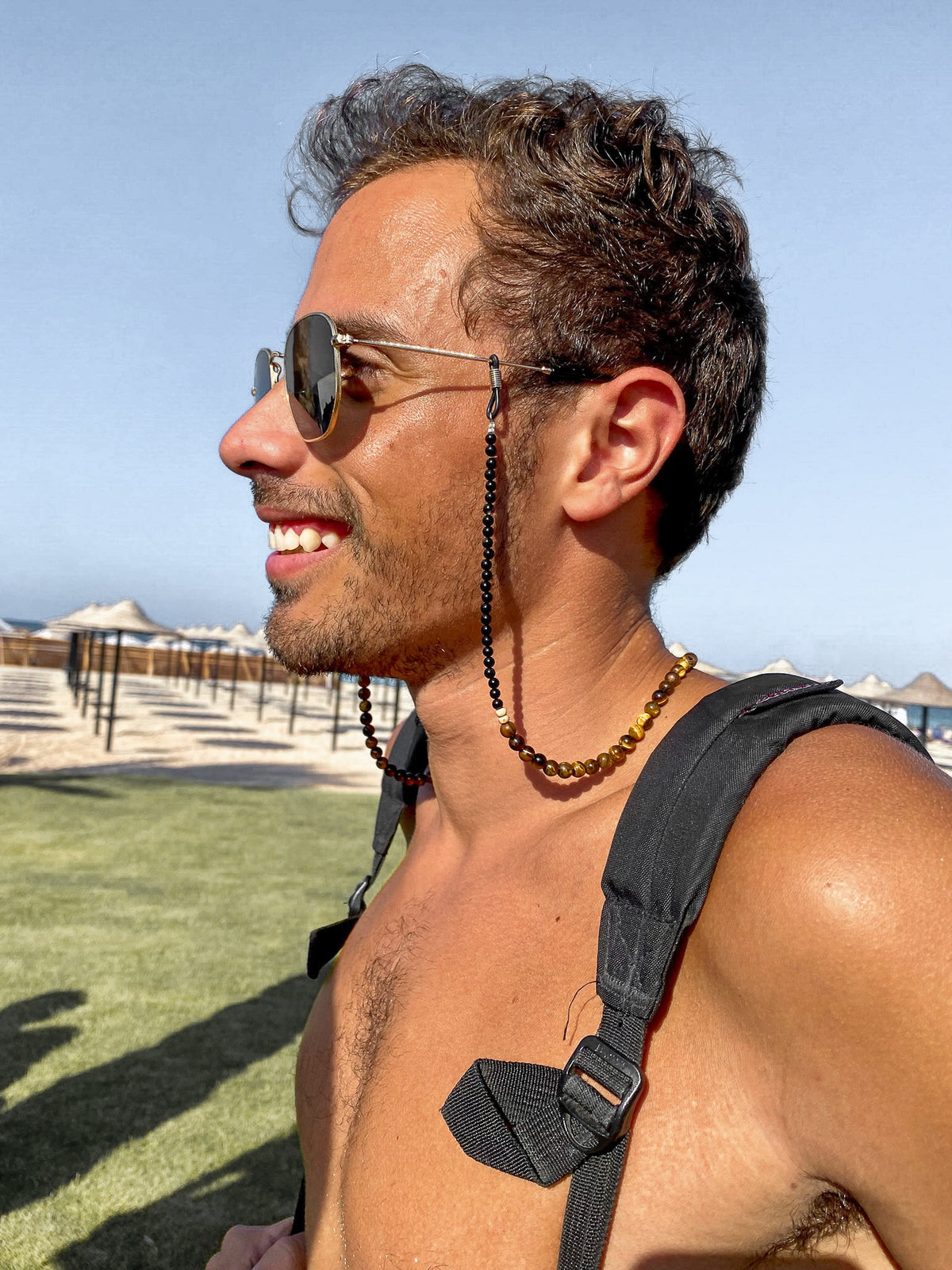 Man with sunglasses and sunglass chain consisting of onyx and tiger eye stones and wearing a backpack standing outdoors on a sunny day.
