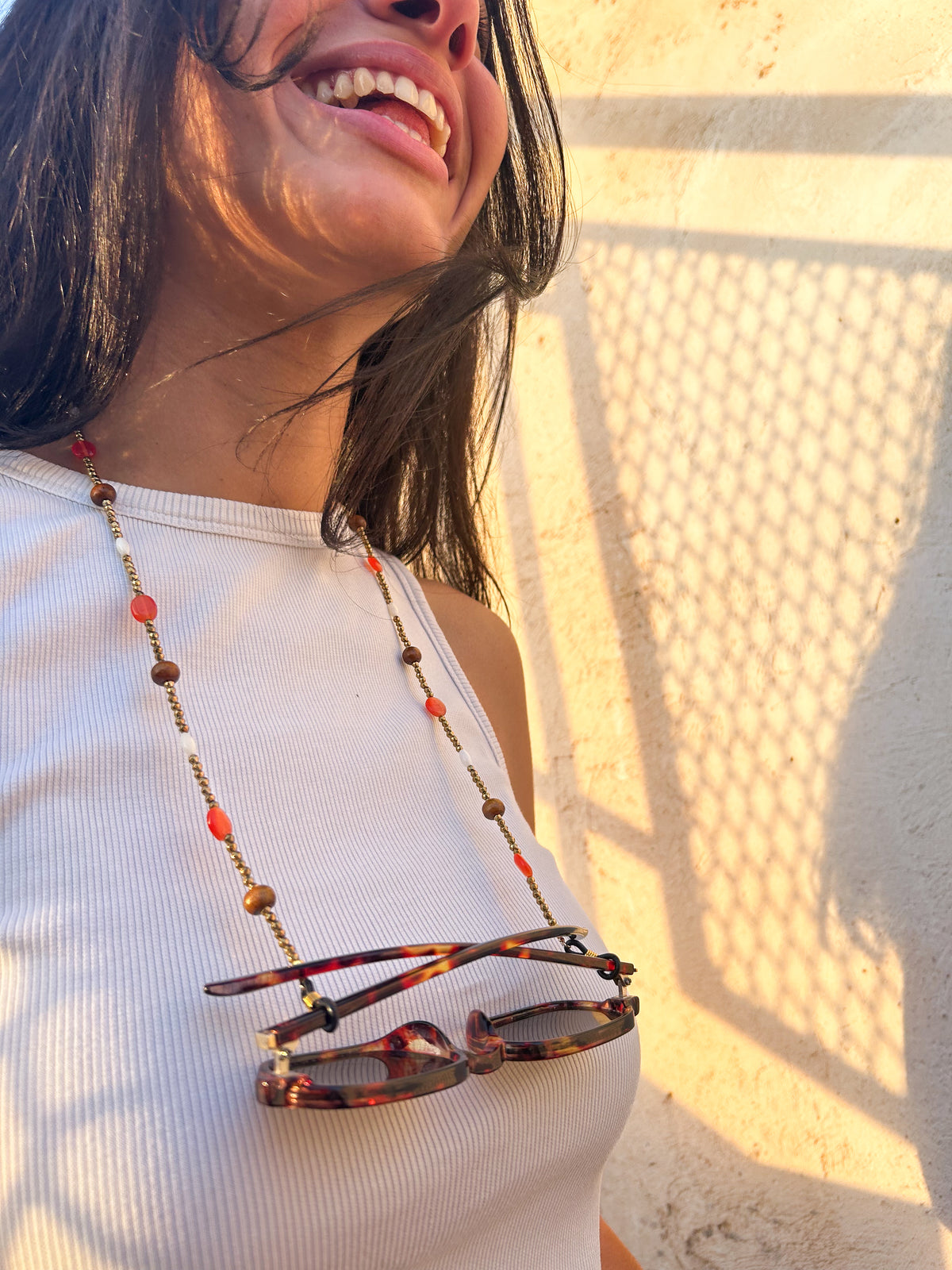 A close-up lifestyle shot of a woman wearing a beaded sunglass chain with warm wooden beads, coral accents, ivory details